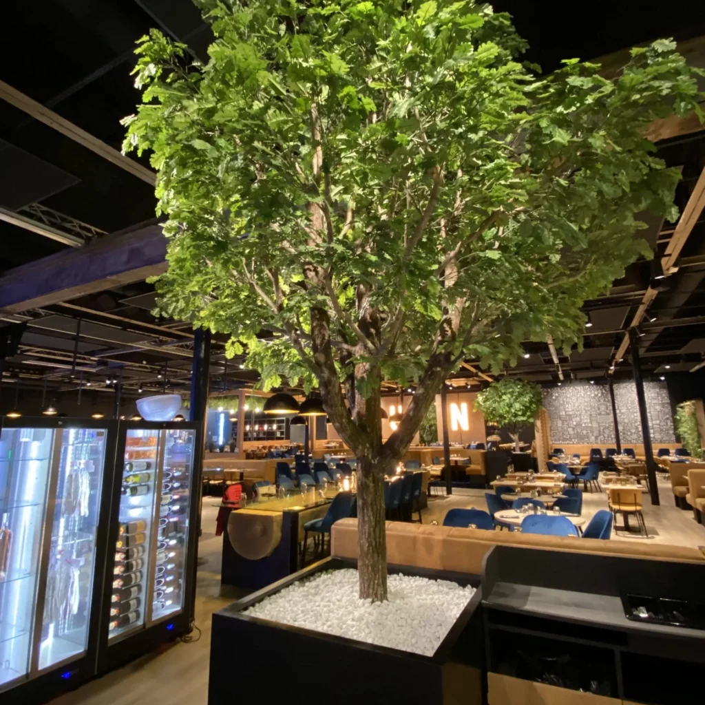 Photo d'un arbre semi-naturel dans un pot rectangulaire noir, recouvert de gravier blanc, devant des banquettes dans une salle de restaurant